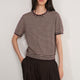 A woman modeling a short-sleeve knit top with horizontal brown and beige stripes, featuring a scalloped neckline and ribbed hem, paired with dark brown pleated trousers, against a plain light gray background.