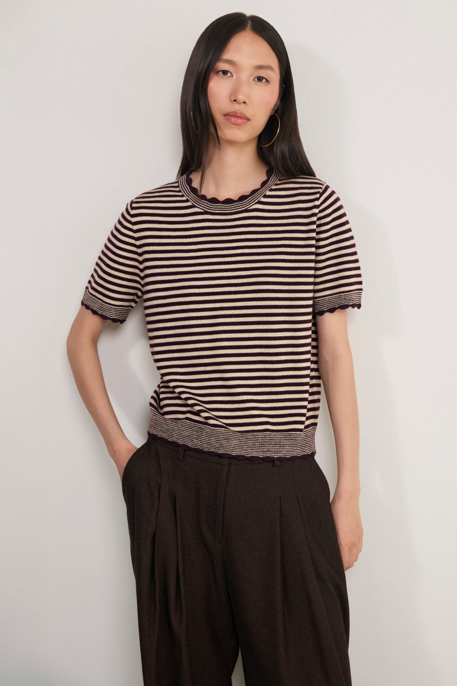 A woman modeling a short-sleeve knit top with horizontal brown and beige stripes, featuring a scalloped neckline and ribbed hem, paired with dark brown pleated trousers, against a plain light gray background.