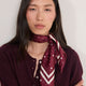 A woman with long dark hair wearing a burgundy and white patterned silk neck scarf tied around her collar, paired with a matching burgundy knit top and gold hoop earrings, set against a plain off-white background.