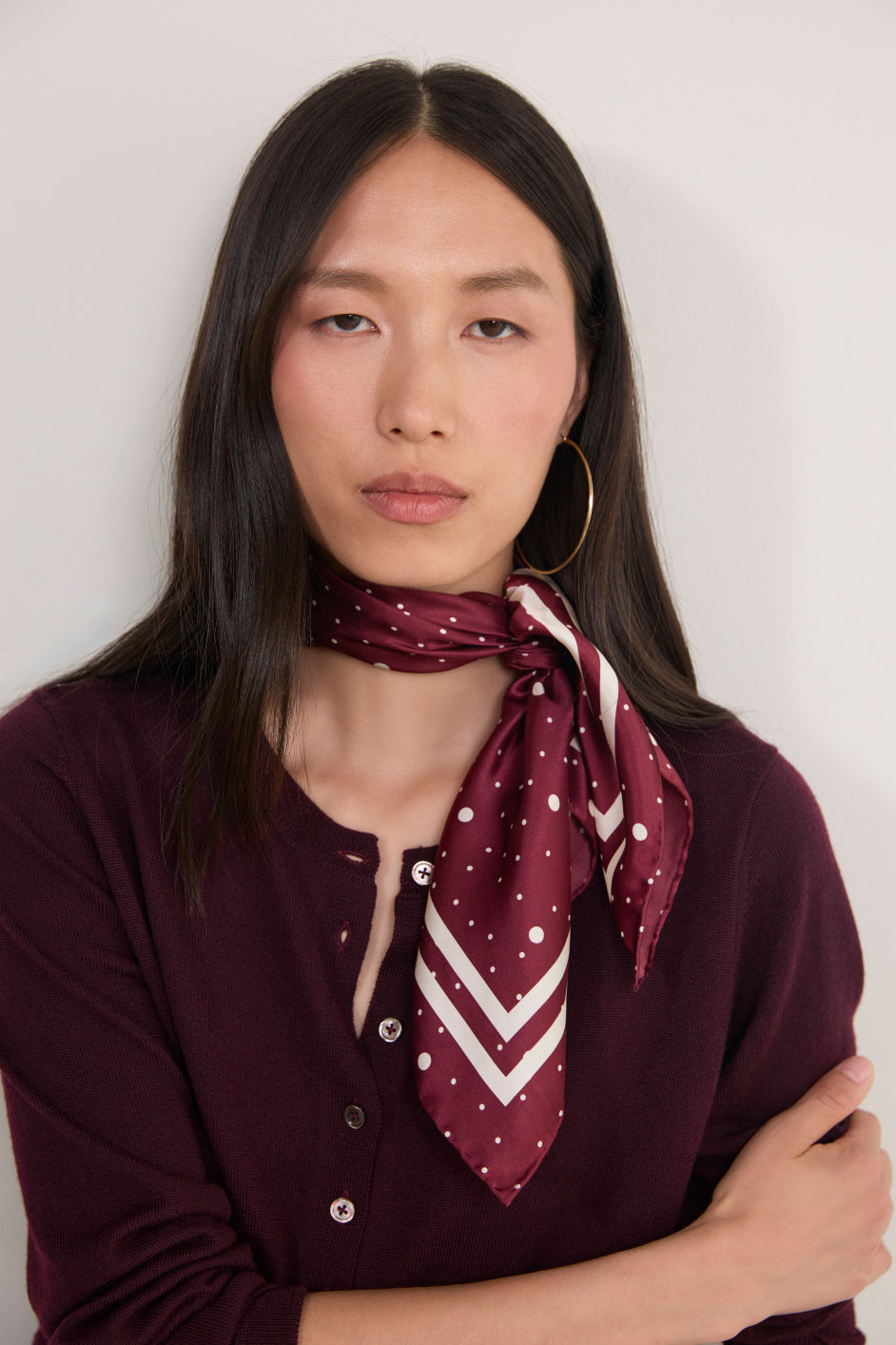 A woman with long dark hair wearing a burgundy and white patterned silk neck scarf tied around her collar, paired with a matching burgundy knit top and gold hoop earrings, set against a plain off-white background.
