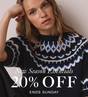 A woman wearing a navy knit sweater with a white and blue Nordic fair isle pattern, posed against a neutral beige wall.